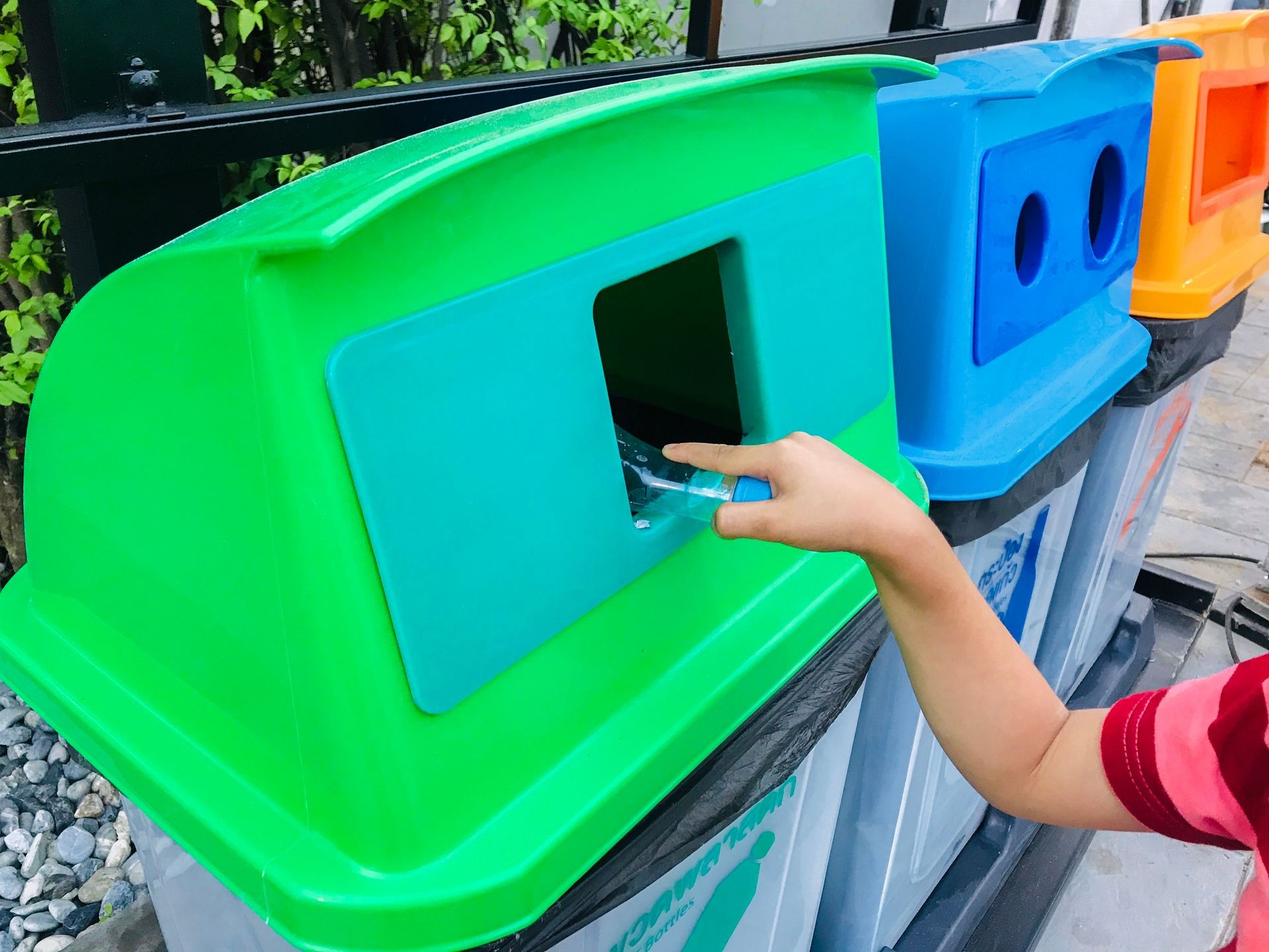 Caucasian hand’s kid putting clear plastic bottle in recycling green bin outside in yard. Close up hand’ boy throwing empty bottle into savings reduce junk. Text on trash meaning Kind of garbage.
